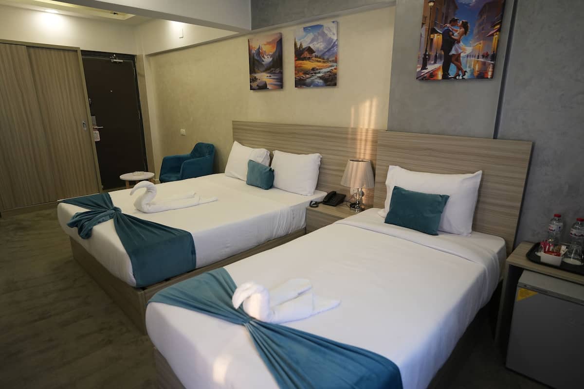 Una habitación de hotel con dos camas, cada una con una toalla azul y almohadas blancas. Hay cuadros en la pared y una mesita con una lámpara y una botella.