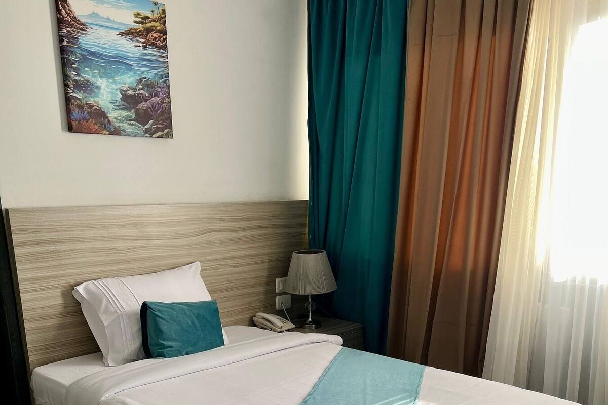 Una cama cuidadosamente hecha con ropa de cama blanca y azul aguamarina, un cabecero de madera, una mesita de noche con una lámpara y un cuadro en la pared.