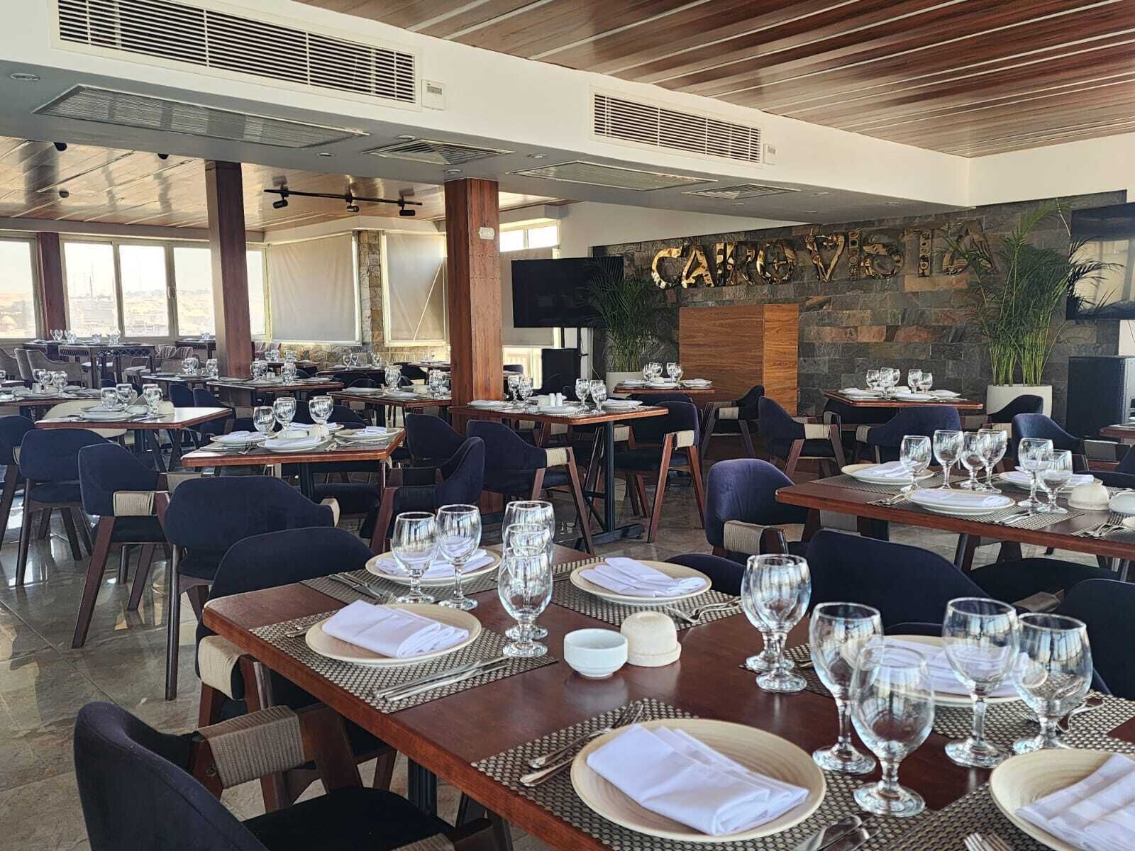 A restaurant with multiple tables set for dining, featuring a stone wall with the name 'CAIRO' and a large screen.