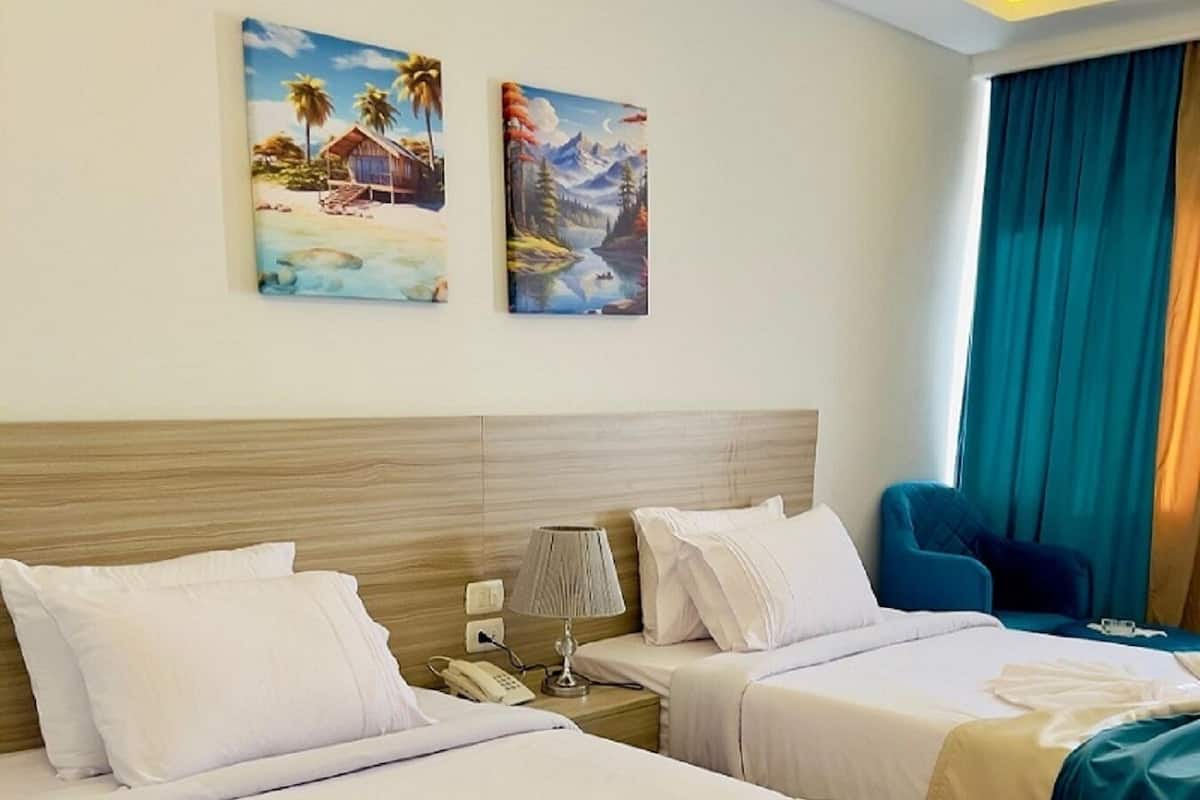 Habitación de hotel con dos camas, una mesita de noche con lámpara, una silla y una obra de arte en la pared.