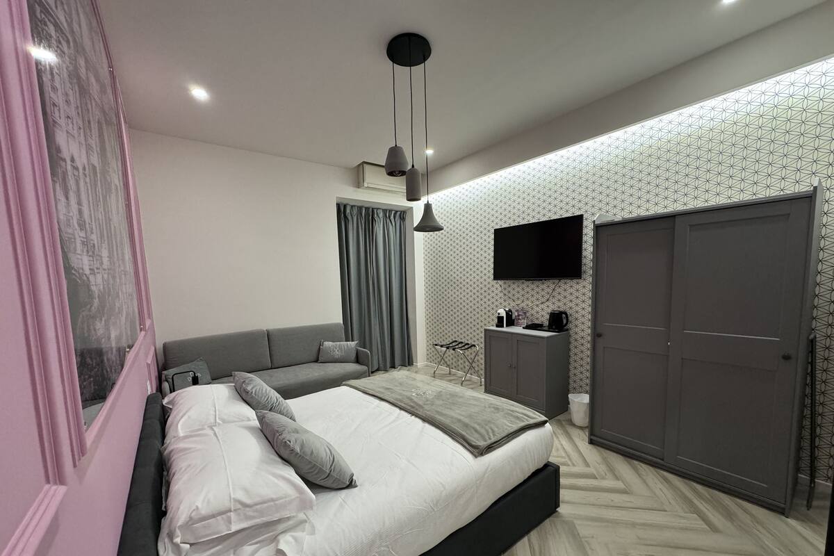 A modern bedroom with a bed, grey sofa, wardrobe, and a TV on the wall.