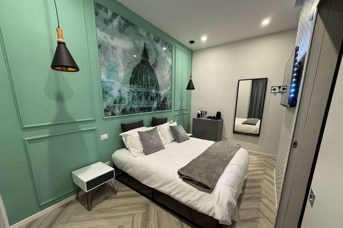 A modern bedroom with a large bed, a nightstand, a small table, a mirror, and a wall-mounted light fixture.
