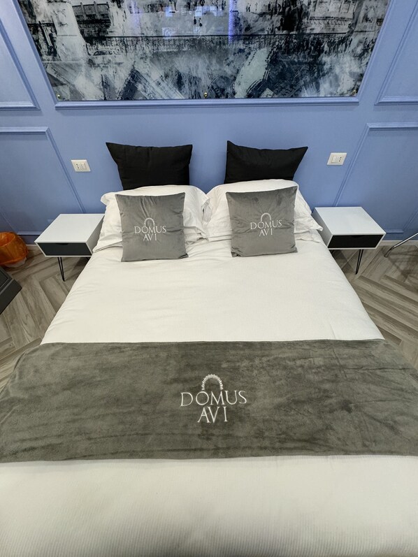 Una cama bien hecha con dos almohadas con la etiqueta "DOMUS AVI" y una manta gris con el mismo logotipo.