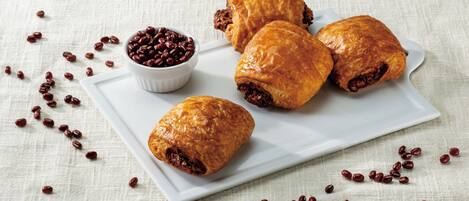 Four pastries on a white plate with a small bowl of beans beside them, scattered beans around the plate, and a textured fabric background.