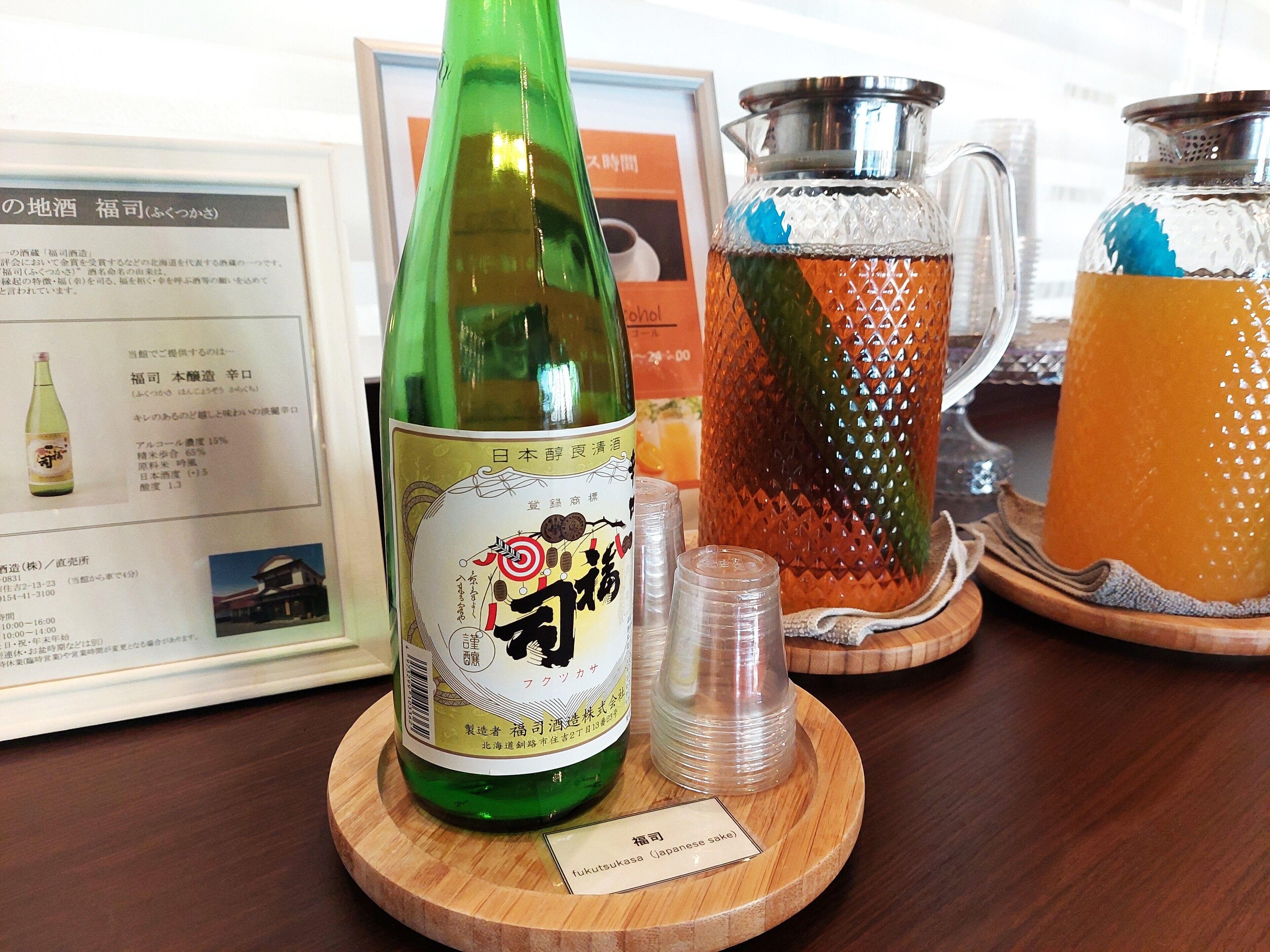 A bottle of sake on a wooden coaster with a label, a glass pitcher with a handle, and a glass with a straw.