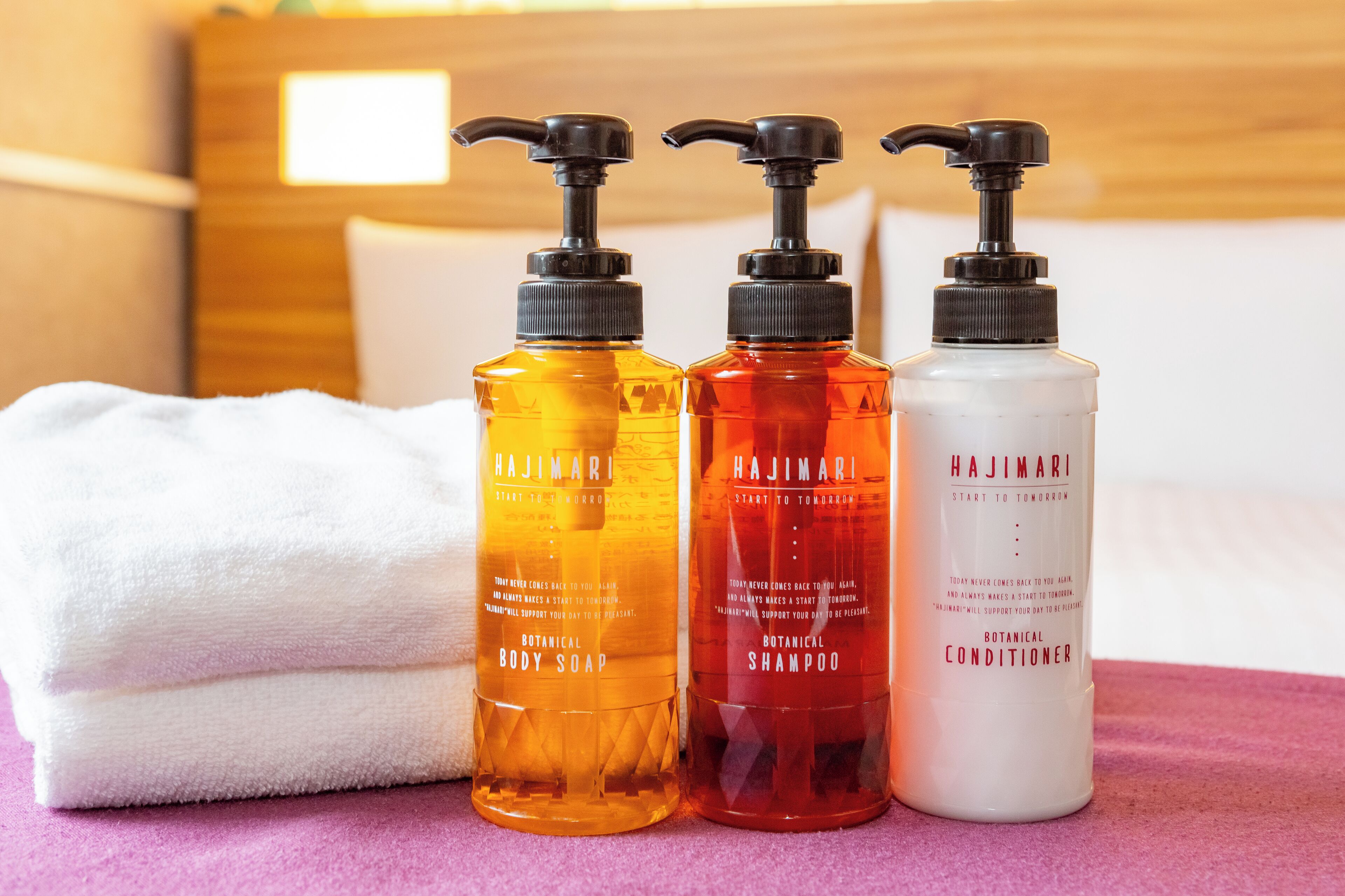 Drie Hajimari producten: douchezeep, shampoo en conditioner, op een roze oppervlak met een witte handdoek en een houten hoofdbord op de achtergrond.
