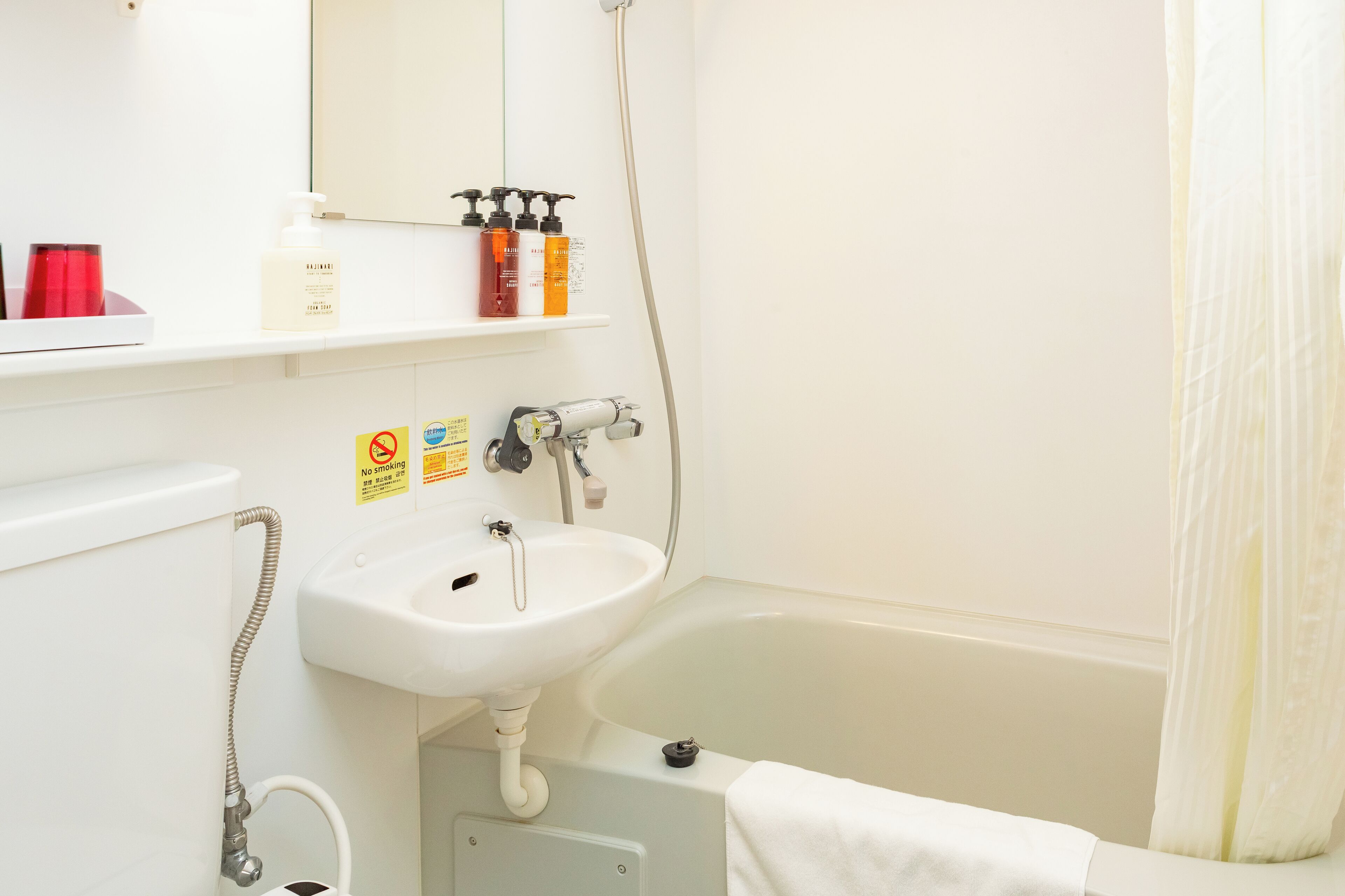 Un baño con lavamanos blanco, bañera y ducha. Hay artículos de aseo en el estante y un cartel de no fumar.