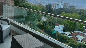 Een balkon met rieten meubilair, een glazen hekwerk en een uitzicht op een stadsbeeld met gebouwen en bergen op de achtergrond.