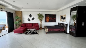 A modern living room with a red sectional sofa, a glass coffee table, a bed with a dark headboard, and a large Buddha painting on the wall.