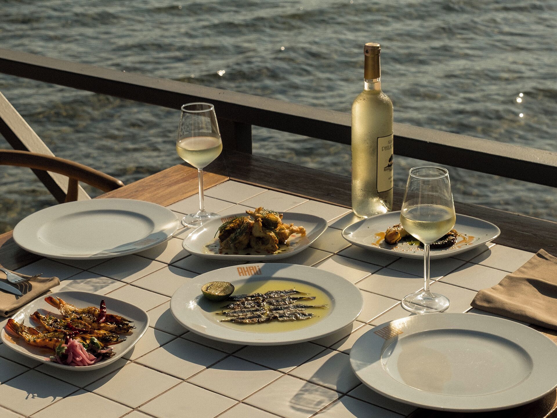 Une table dressée avec des assiettes remplies de nourriture, des verres de vin et une bouteille de vin, donnant sur une étendue d’eau.