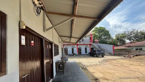 A covered outdoor area with a paved walkway, a dark brown door, and a building with multiple doors in the background. - Steze Guesthouse Syariah Telanaipura (Jambi)