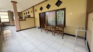 A spacious room with a tiled floor, a wooden dining table with chairs, a window with curtains, and a small white stand. - Taxa Raya Guest House (Kuta)