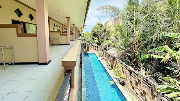A pool area with a tiled floor, a long bench, and a stone wall with tropical plants. - Taxa Raya Guest House (Kuta)