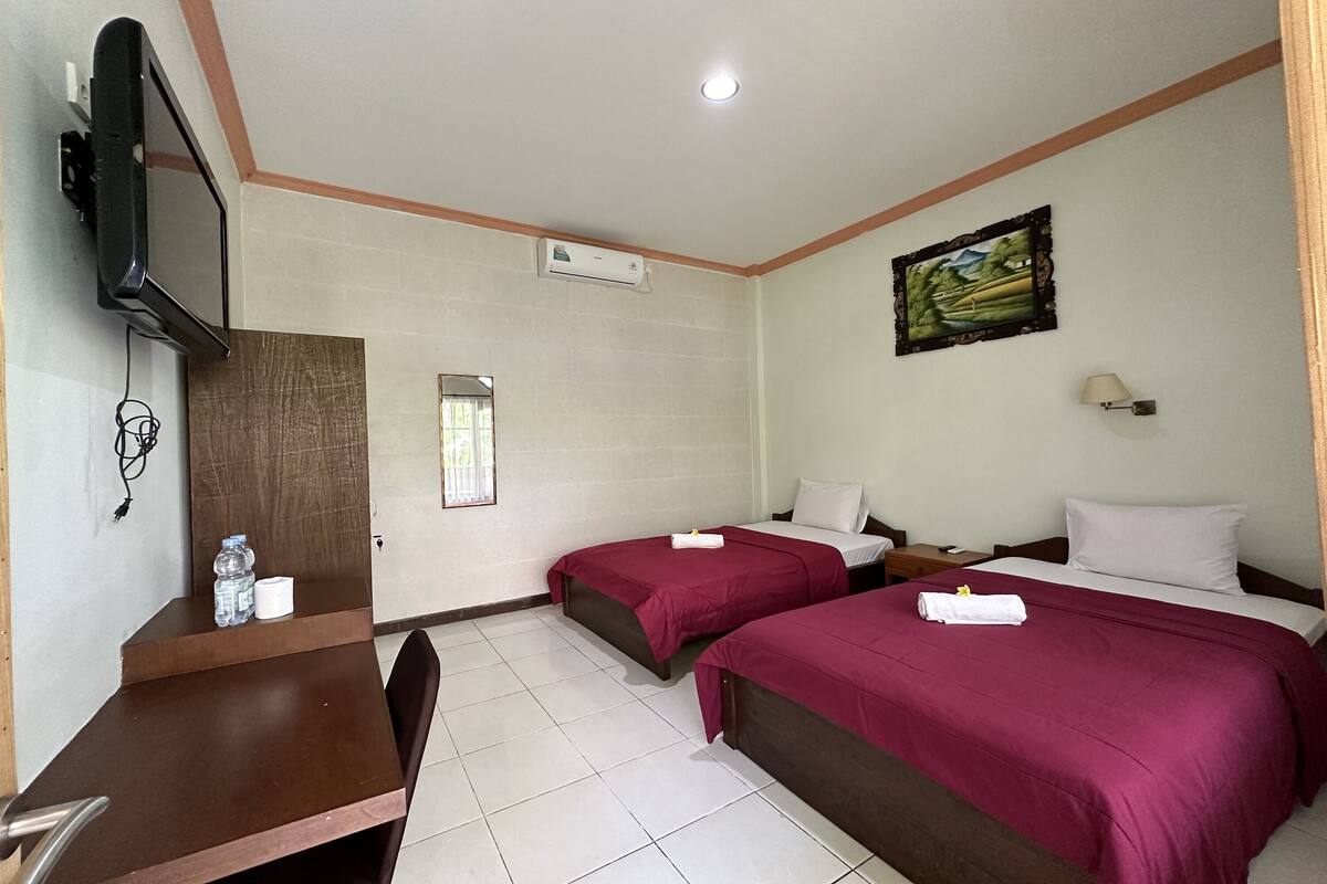 A hotel room with two beds, a desk, a TV, an air conditioning unit, and a painting on the wall.