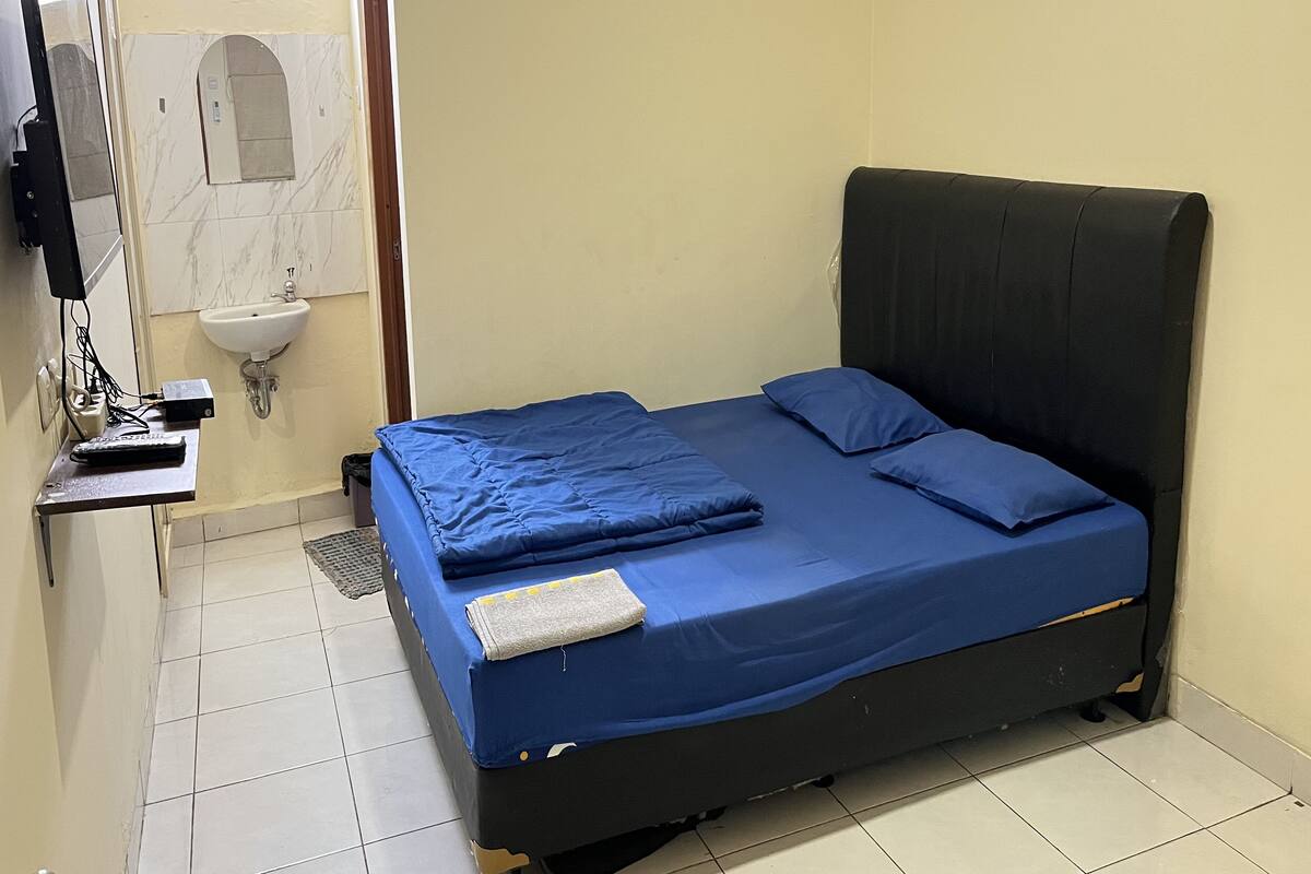 A small, single-bed room with a black headboard, a blue bedspread, and a small bedside table with a remote control.