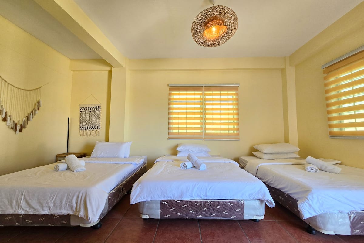 A hotel room with three single beds, a large window with blinds, a hanging light fixture, and a wall decoration.