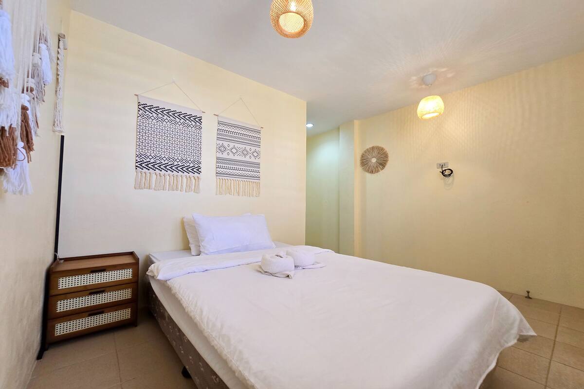 A neatly made bed with white linens and towels, a wall-mounted macrame piece, a small wooden nightstand, and a wall light fixture.