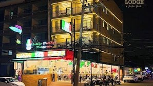A multi-story building with a brightly lit restaurant on the ground floor, a visible sign for a hotel, and several parked vehicles. - The Lodge Jomtien 7 (Pattaya)