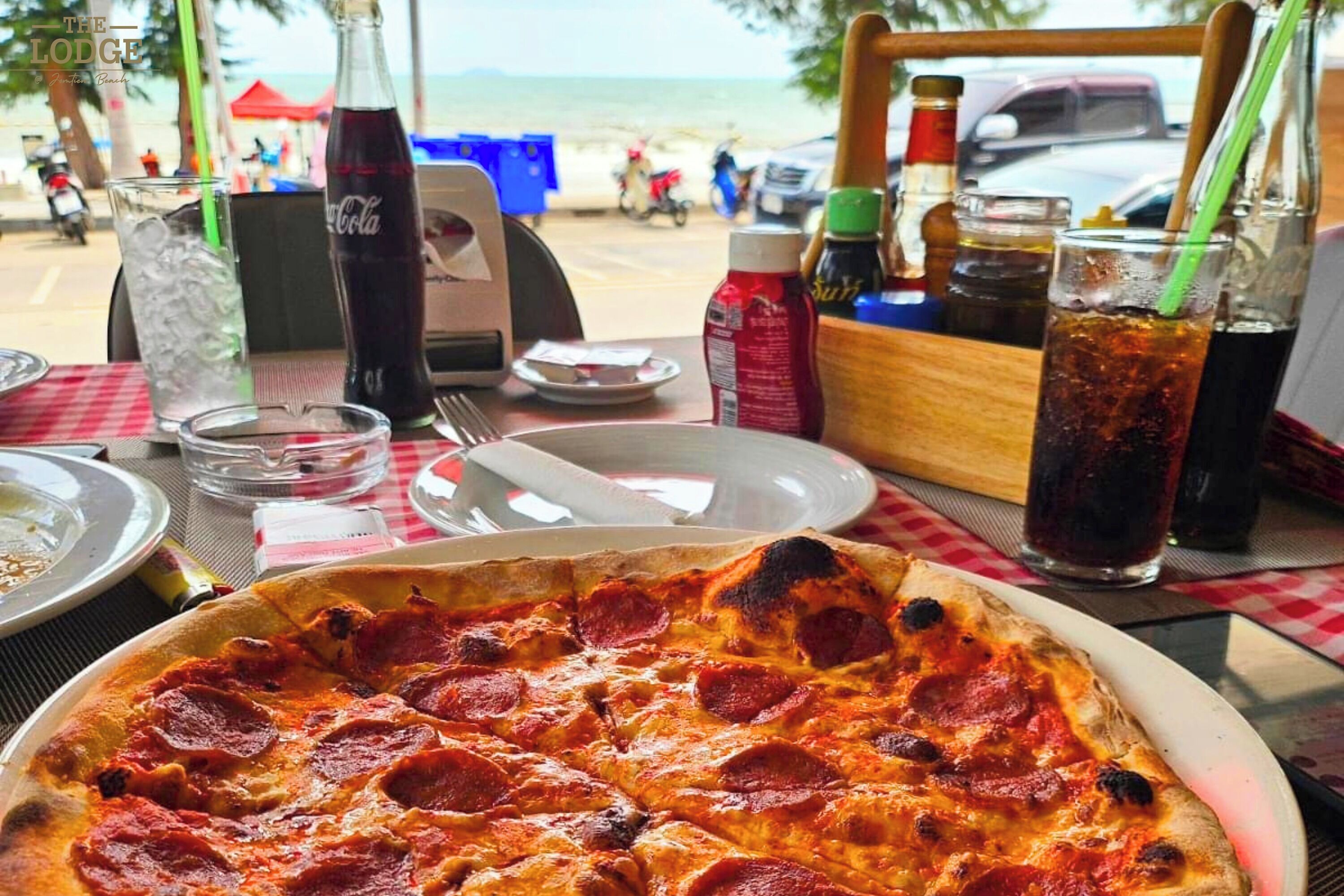 A table with a pepperoni pizza, a Coca-Cola bottle, and a glass of soda at a beachside restaurant.