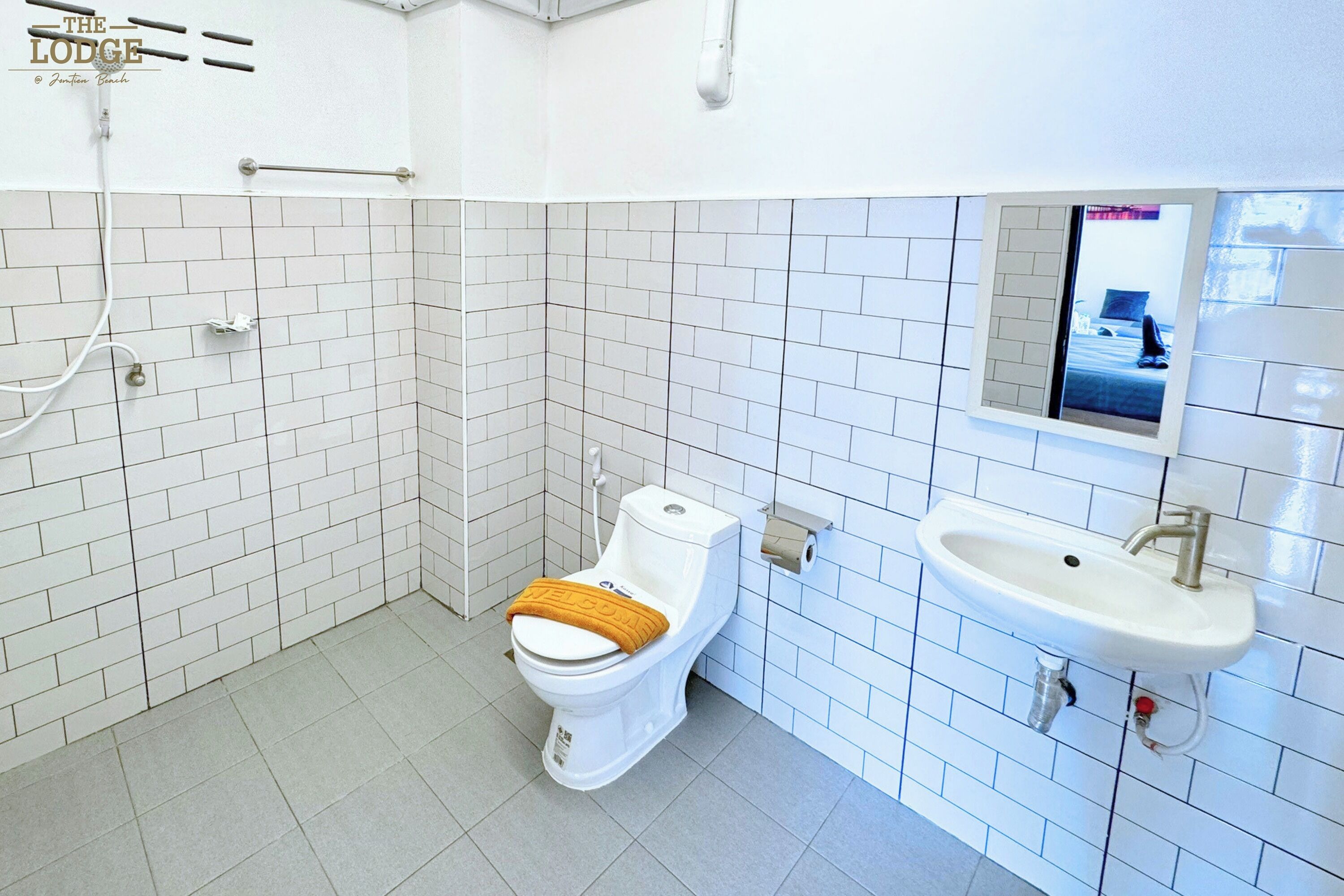 A modern bathroom with white tiled walls, a toilet with a brown seat cover, a sink with a mirror above it, and a window showing a bed.