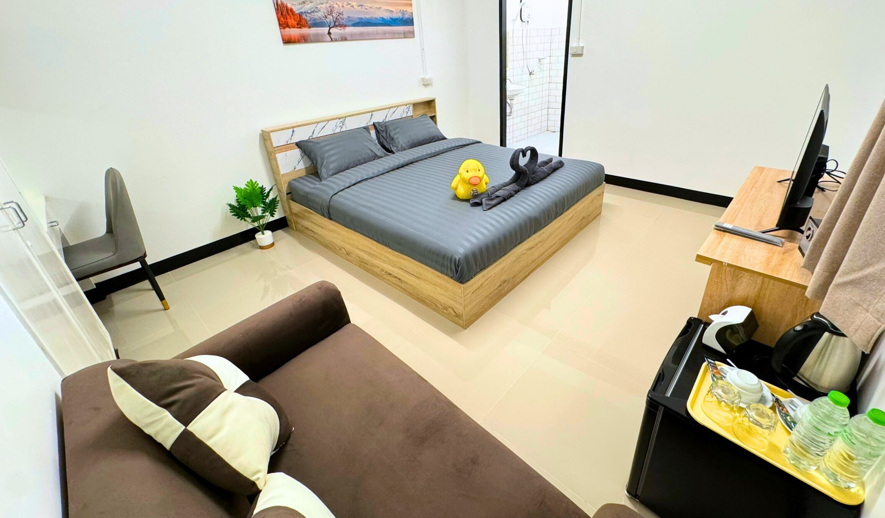 A modern hotel room with a bed, a sofa, a desk with a computer, and a small plant.