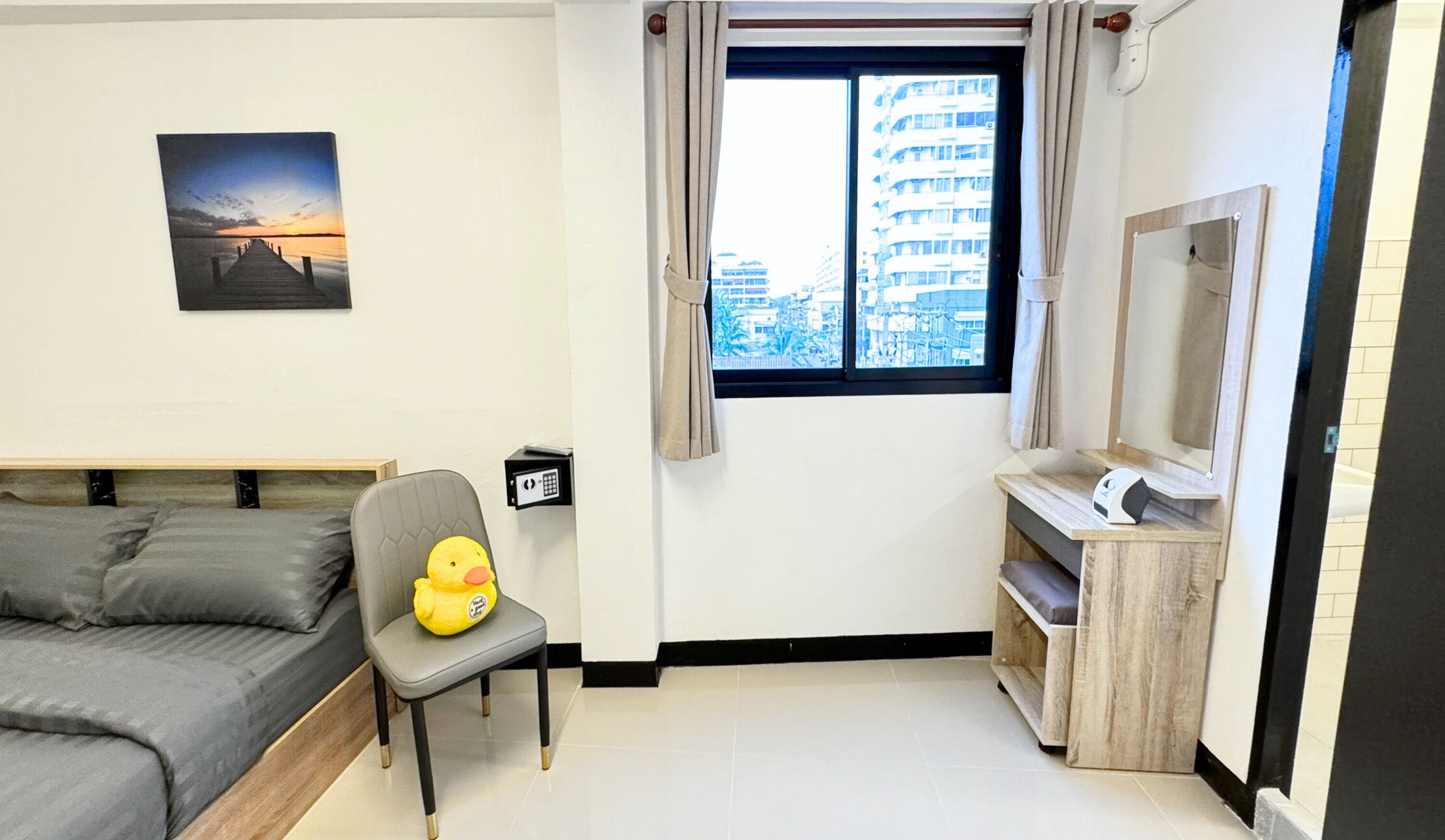 A bedroom with a bed, a chair with a plush toy, a window with curtains, a wall-mounted picture, and a small desk with a mirror.