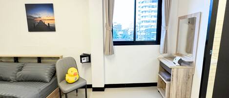 A bedroom with a bed, a chair with a plush toy, a window with curtains, a wall-mounted picture, and a small desk with a mirror.