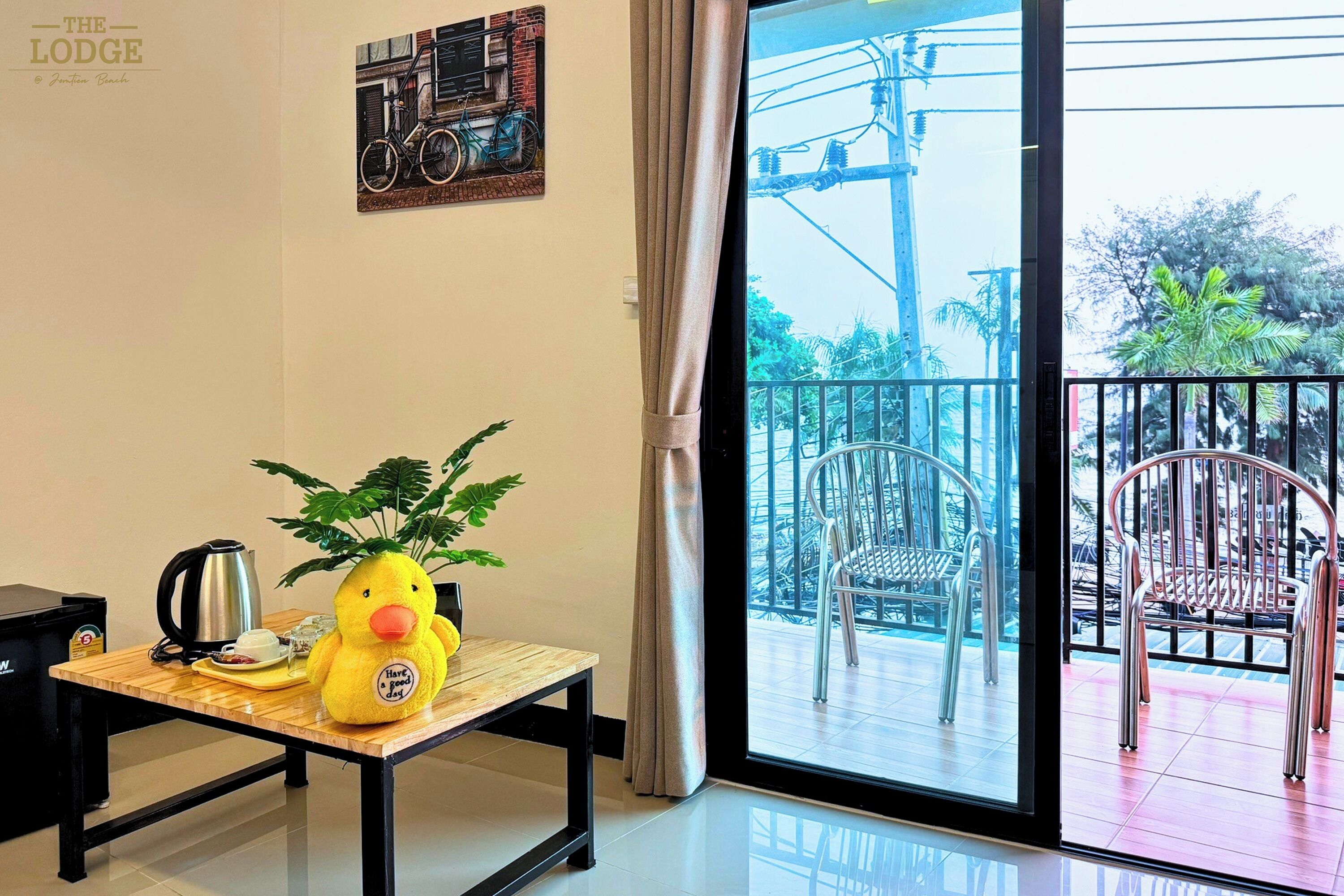 A room with a wooden table, a yellow duck plush toy, a potted plant, a kettle, and a coffee cup. There is a sliding glass door leading to an outdoor balcony with chairs and a railing.