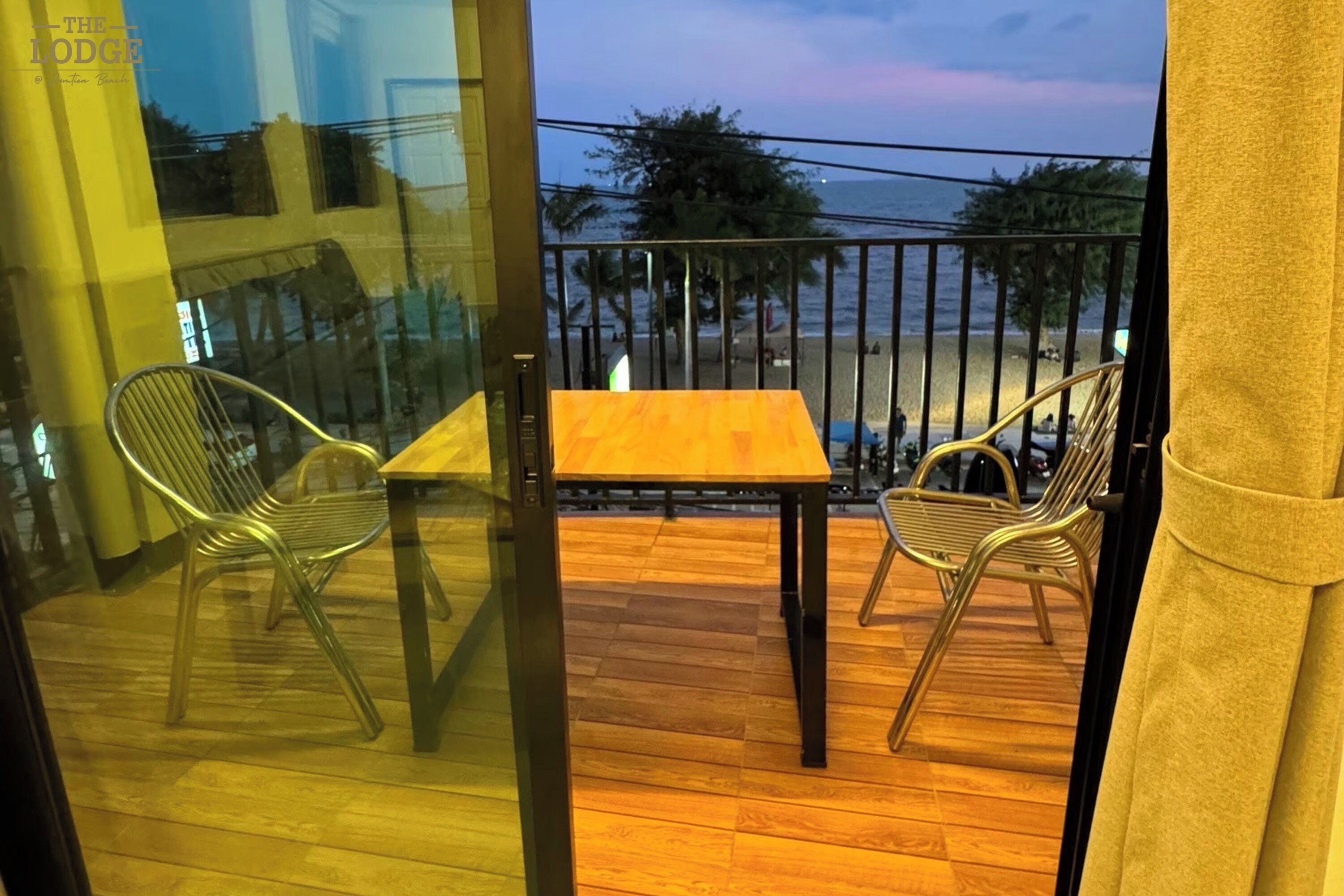 A balcony with a wooden table and two chairs, overlooking a beach with palm trees and a railing.