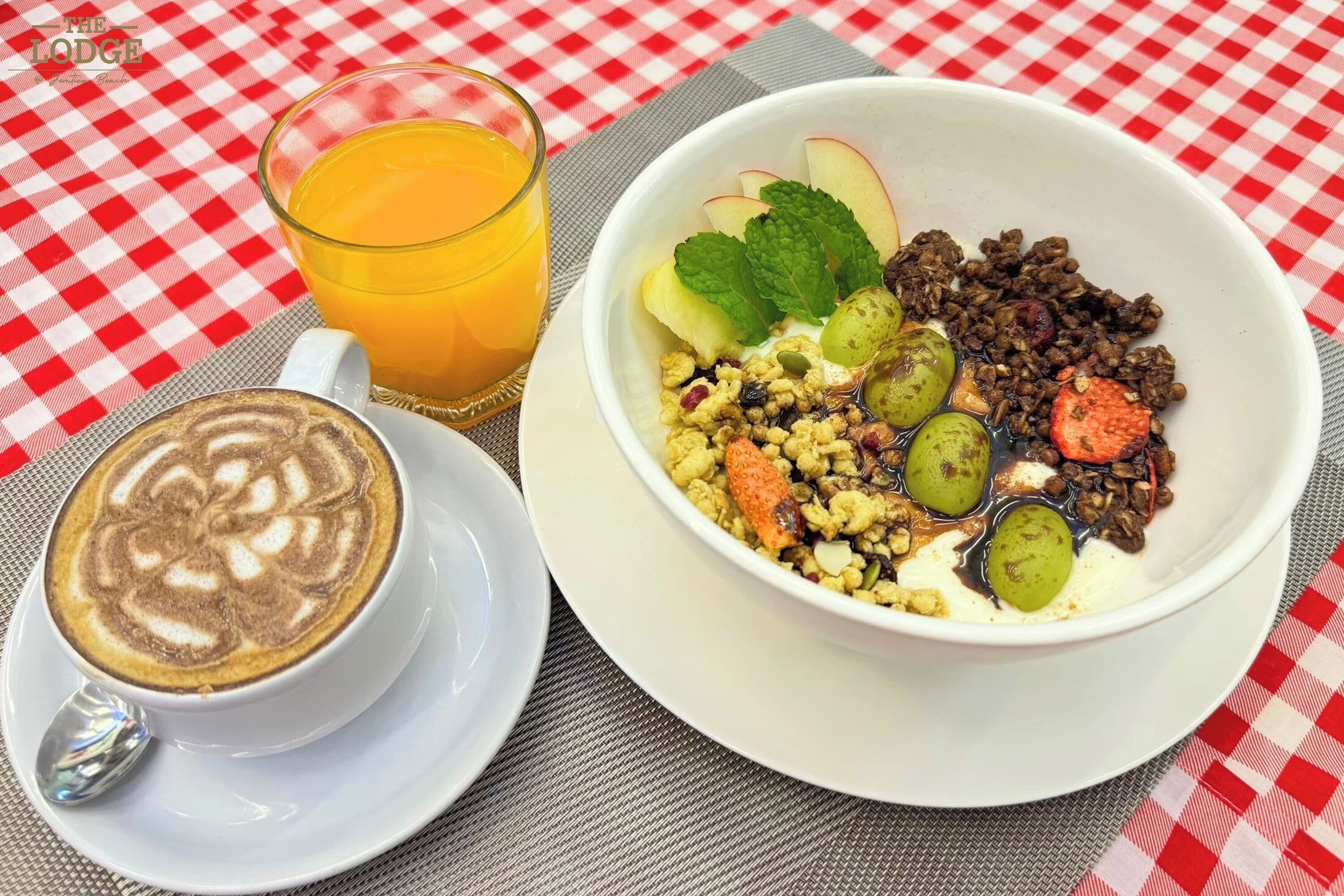 A breakfast setting with a latte, a bowl of cereal with fruits and nuts, and a glass of orange juice on a checkered tablecloth.