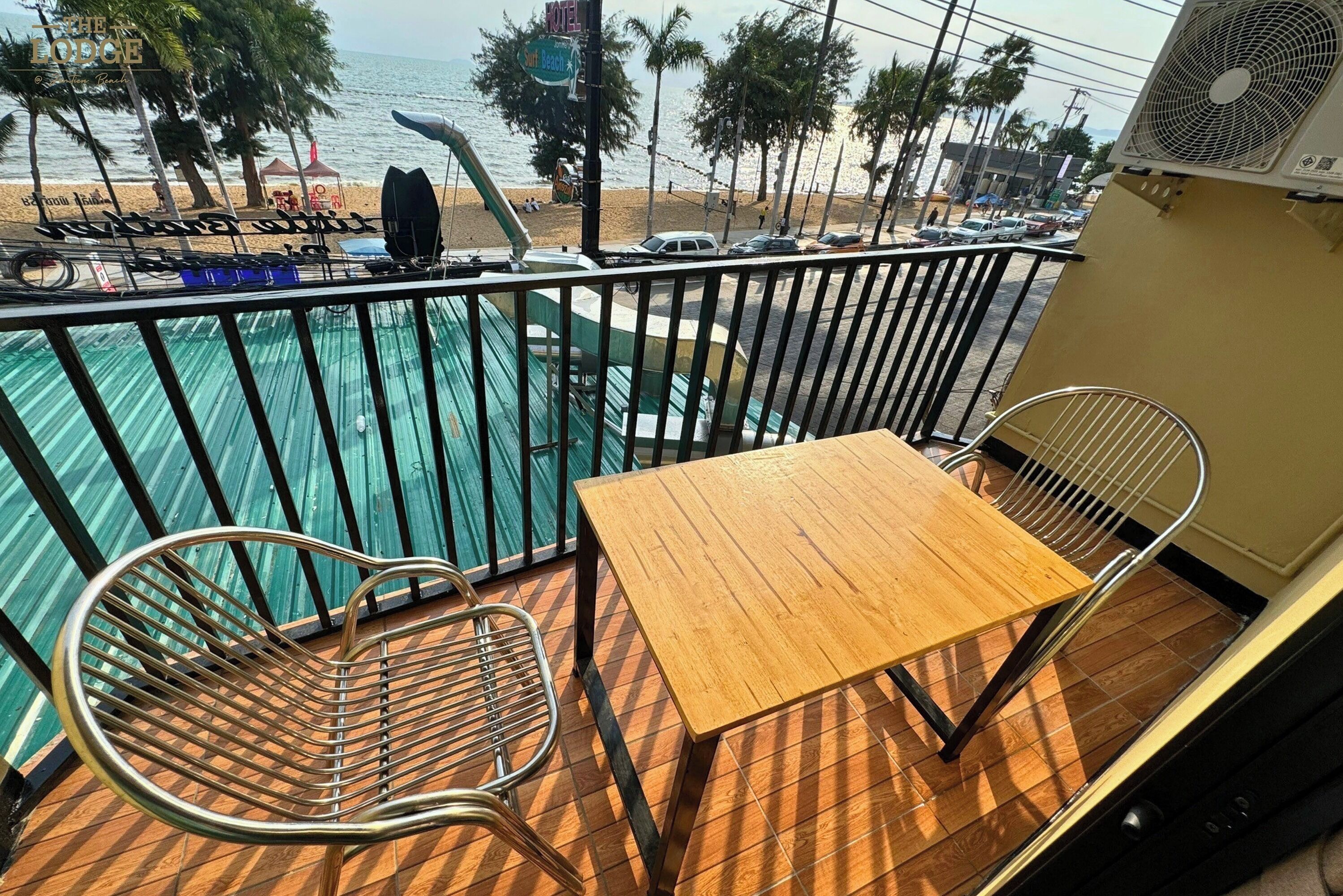 Un balcone con un tavolo di legno e due sedie di metallo, vista su una piscina e una spiaggia con palme e auto parcheggiate.