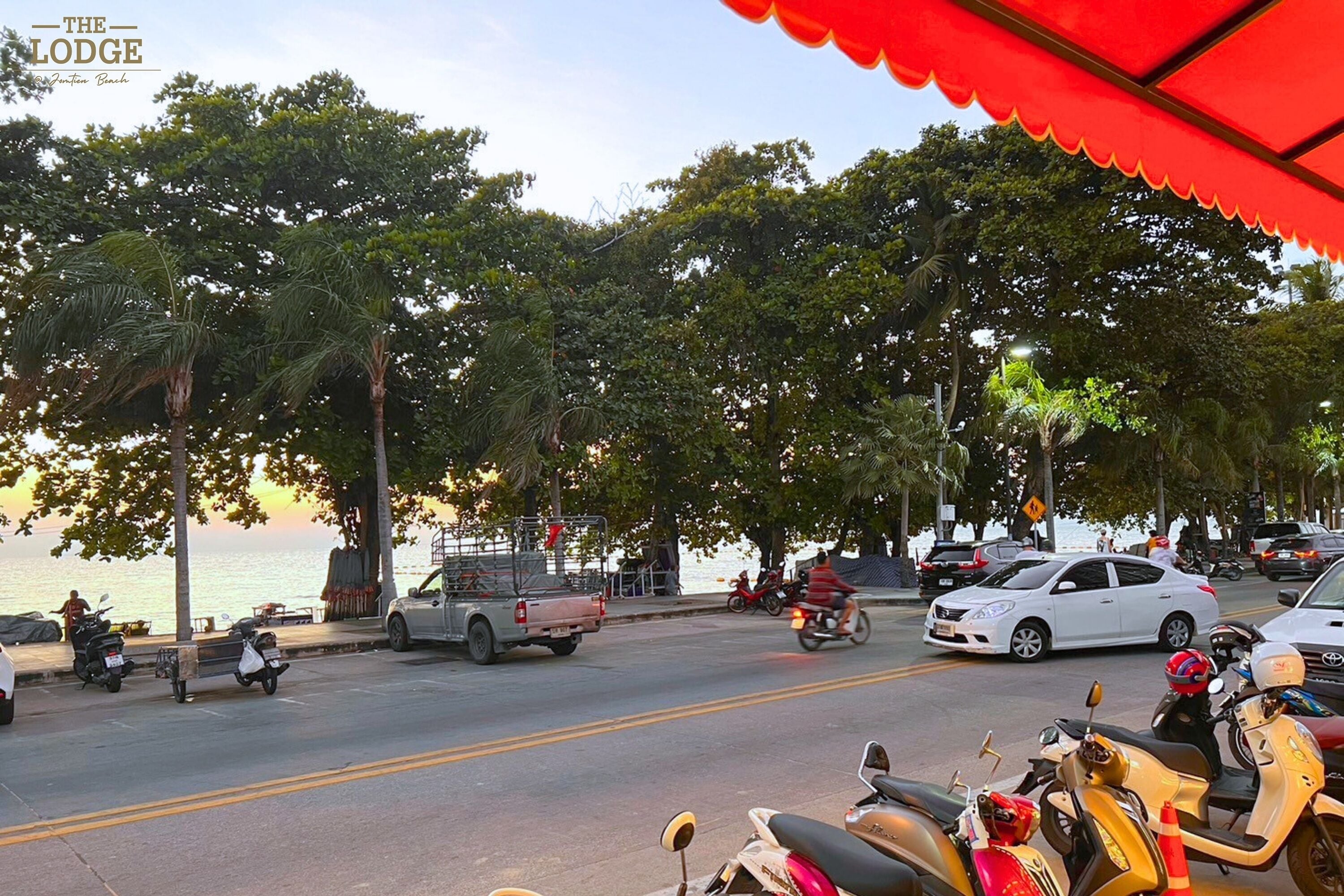 Vista da rua com veículos estacionados, incluindo carros e motocicletas, e algumas pessoas caminhando.