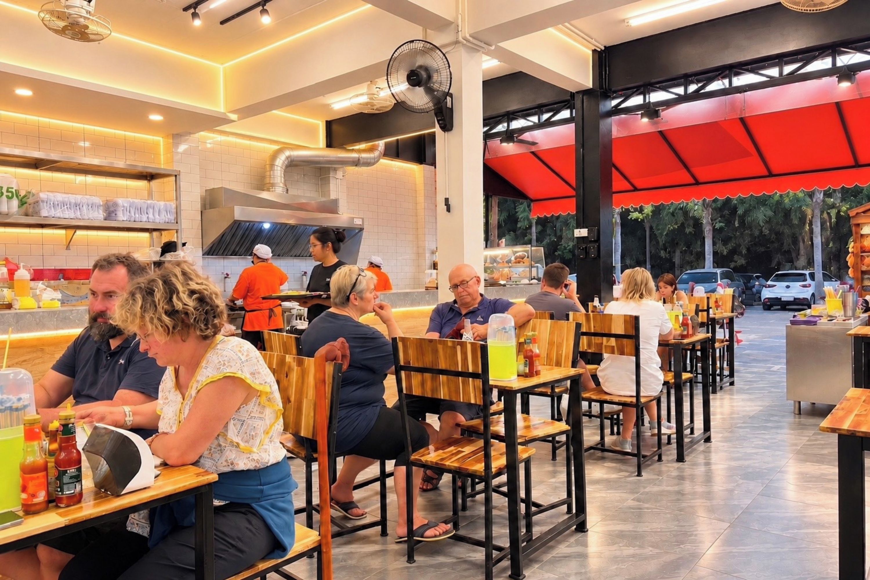 A restaurant with wooden tables and chairs, a kitchen area in the background, and a red awning over the seating area.