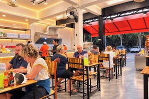 Ein Restaurant mit Holztischen und -stühlen, einem Küchenbereich im Hintergrund und einem roten Markisenvorzelt über dem Sitzbereich.