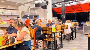 Un restaurant avec des tables et des chaises en bois, un comptoir où sont exposés des plats, et une cuisine visible au fond de la salle.
