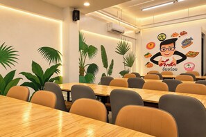 Sala de conferência com mesas e cadeiras de madeira, parede com um mural de comida e um personagem de desenho animado, e um aparelho de ar condicionado.