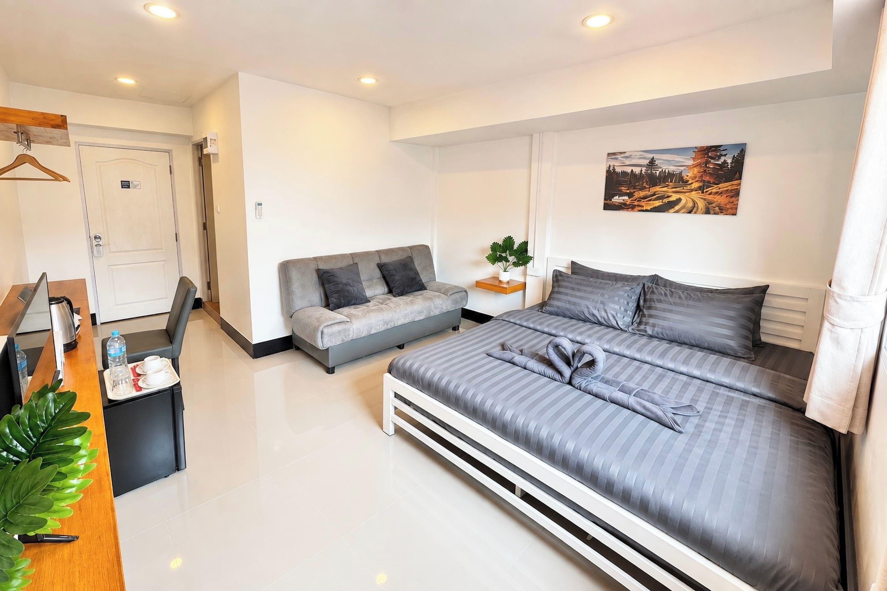 A modern hotel room with a bed, a sofa, a desk with a coffee maker, and a painting on the wall.