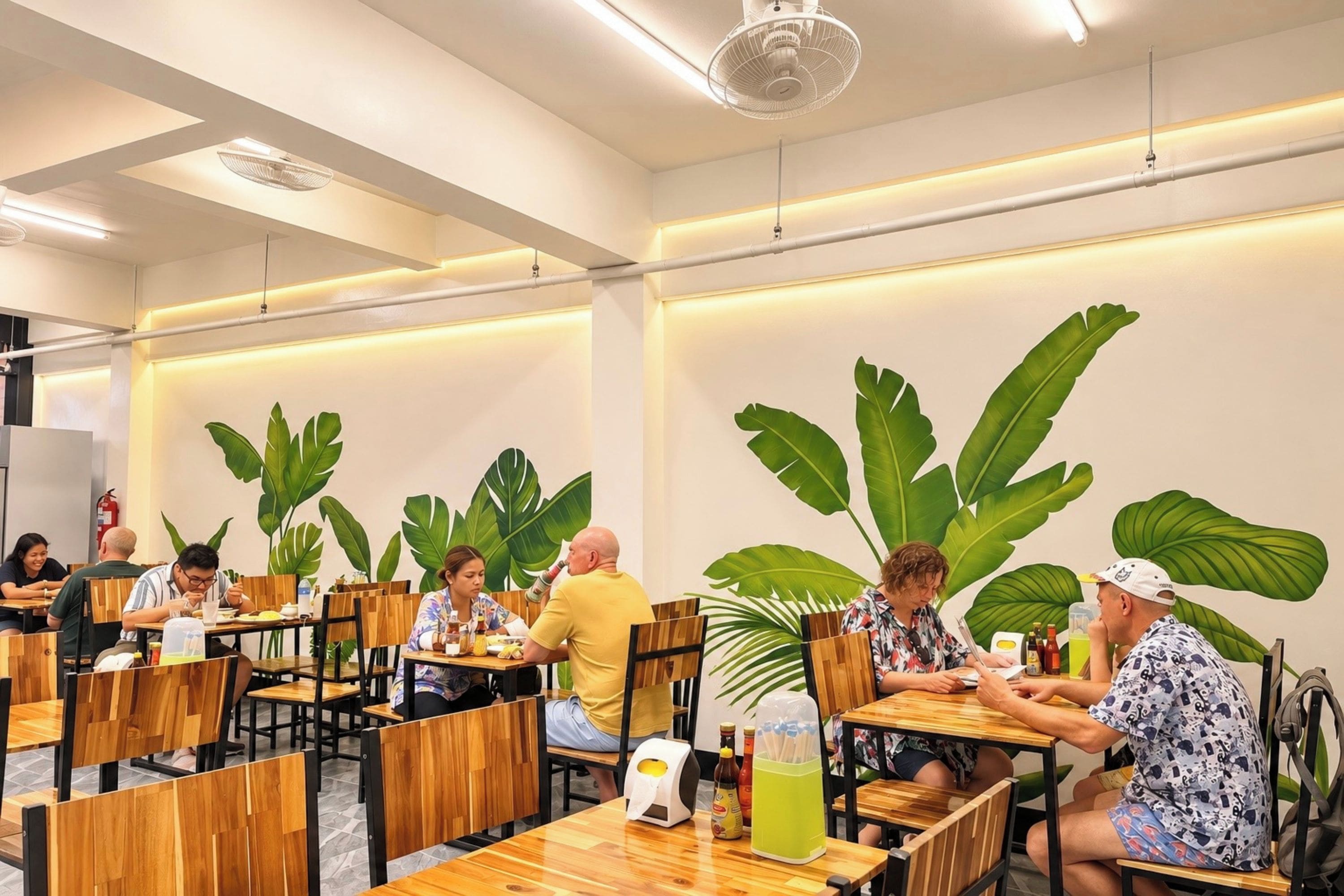 A restaurant with wooden tables and chairs, large green leaves on the wall, and ceiling fans.