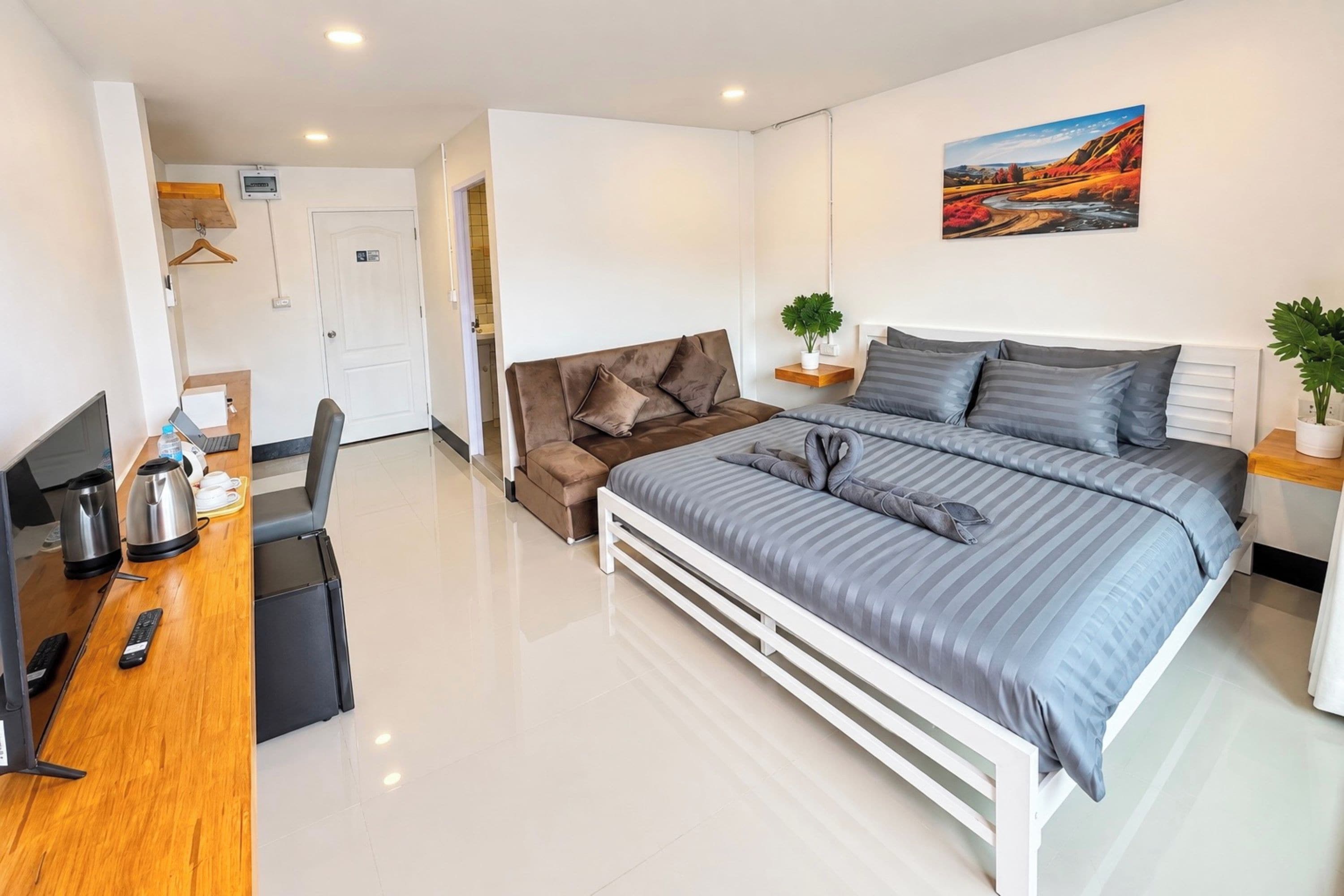 Quarto de hotel moderno com uma cama, uma escrivaninha com um computador, uma cadeira, uma cafeteira e uma pequena planta.