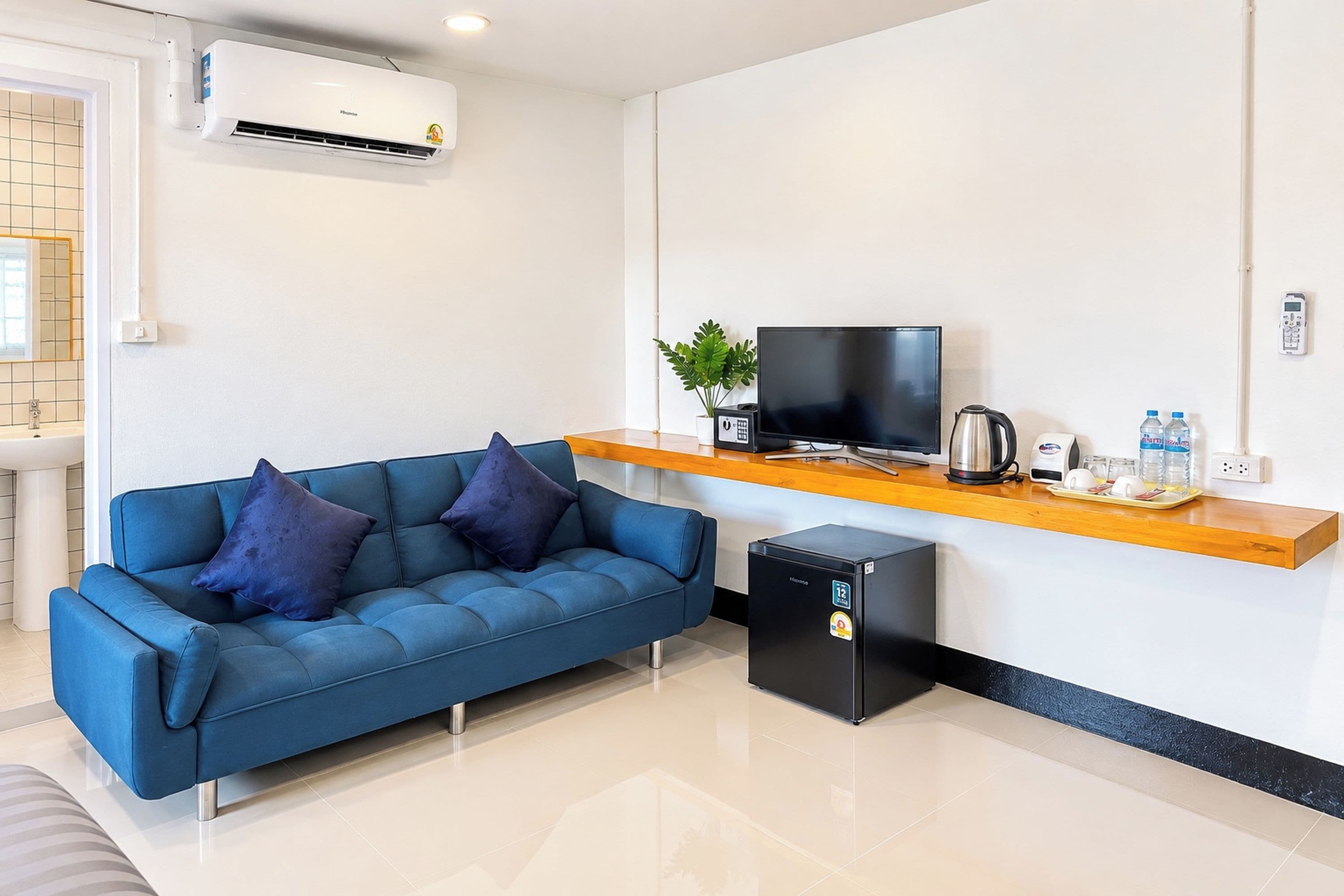 A modern living area with a blue sofa, a small black refrigerator, a television on a wooden shelf, and a coffee maker.