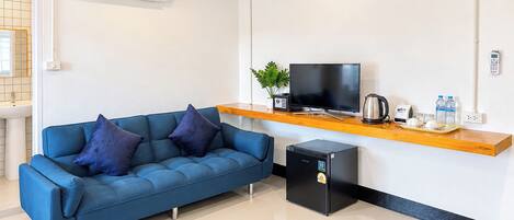 A modern living area with a blue sofa, a small black refrigerator, a television on a wooden shelf, and a coffee maker.