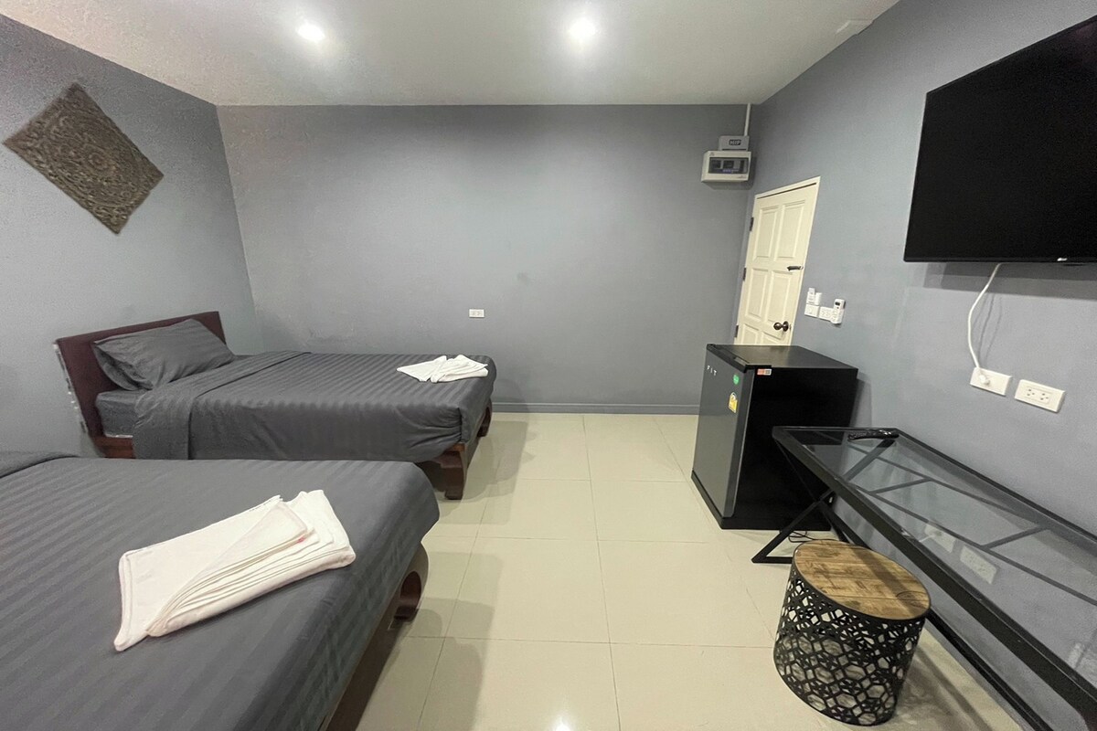 A small, modern room with two single beds, a TV mounted on the wall, a small refrigerator, and a glass table.