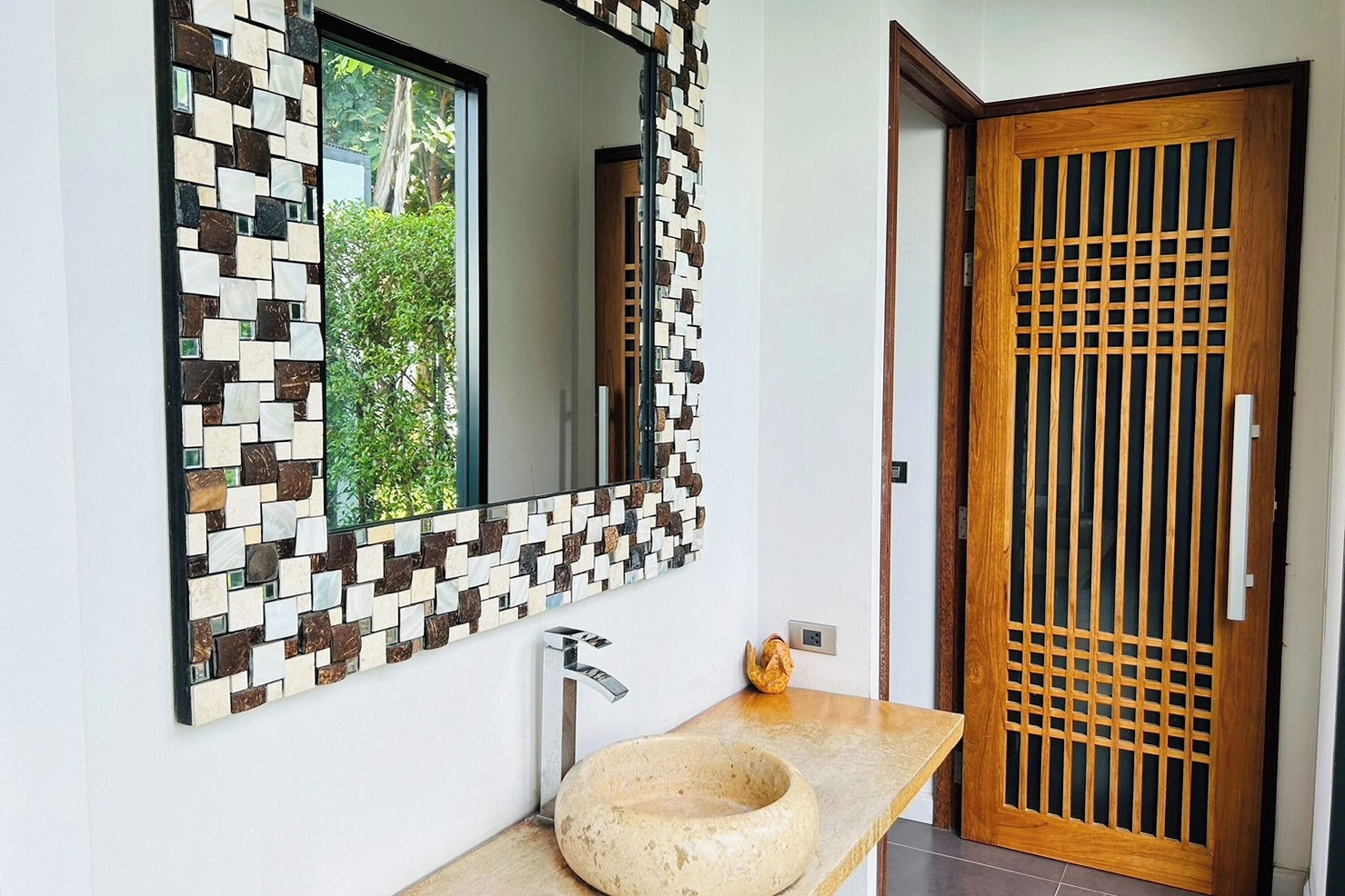 A modern bathroom with a mosaic tile wall, a round sink, and a wooden door with a grid pattern.