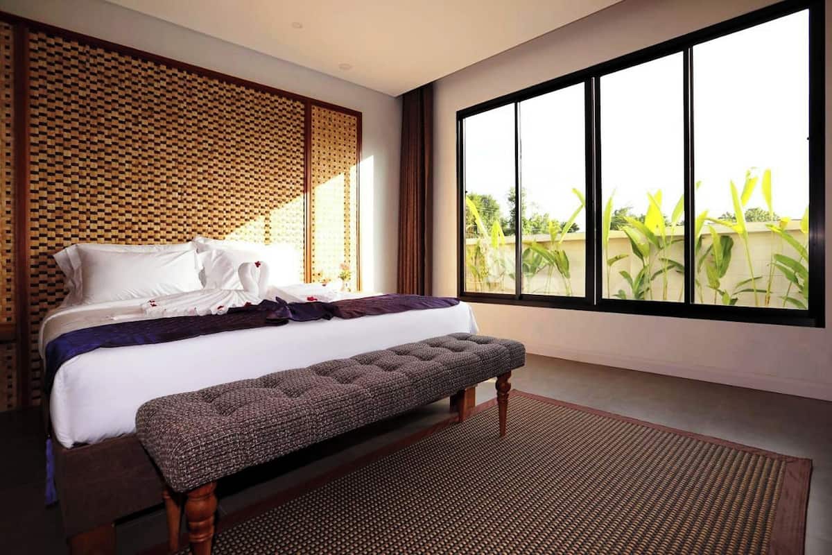 A bedroom with a large window, a bed with white linens, a bench, and a woven wall panel.