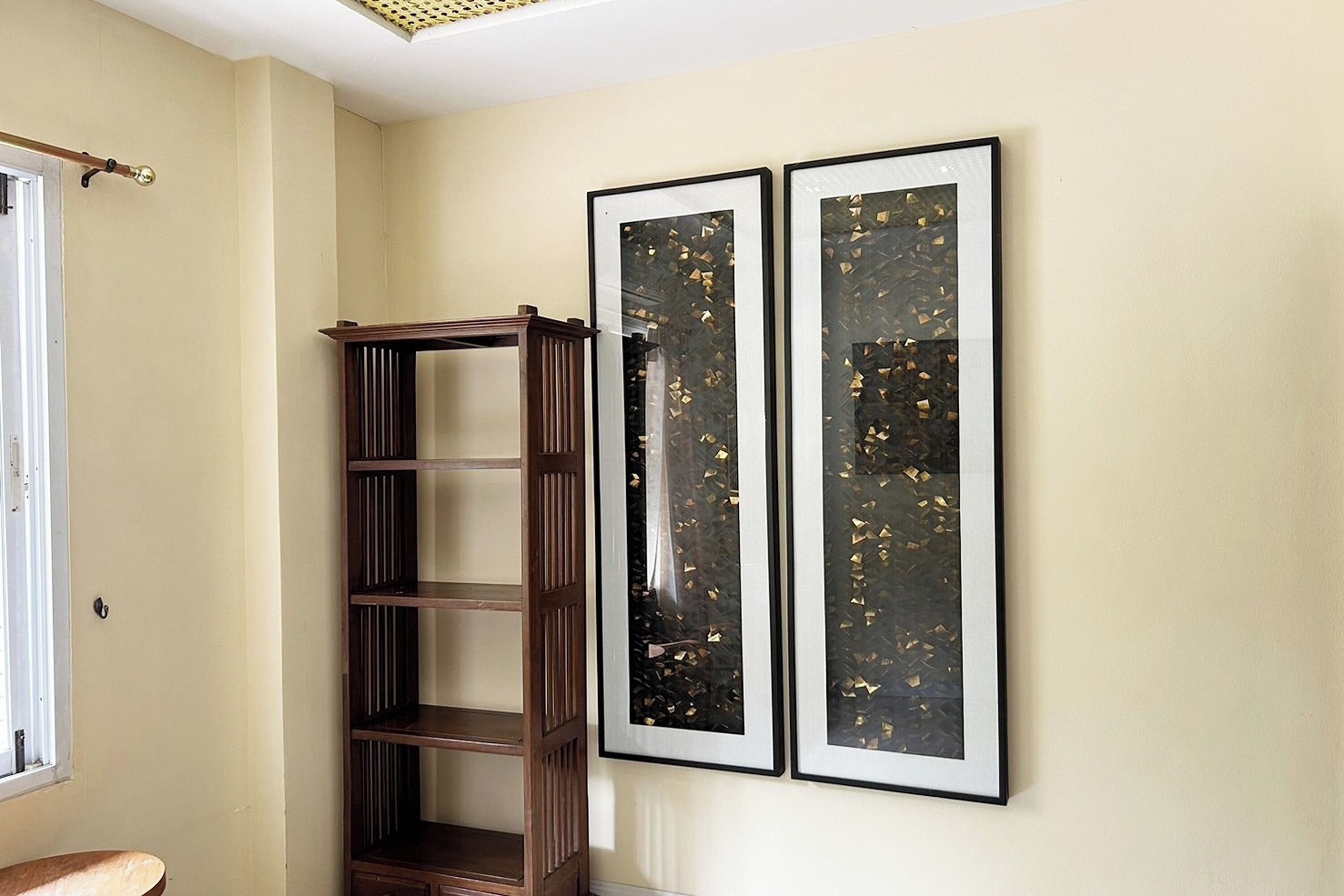 A room with a wooden bookshelf, two framed artworks on the wall, and a window with a curtain.