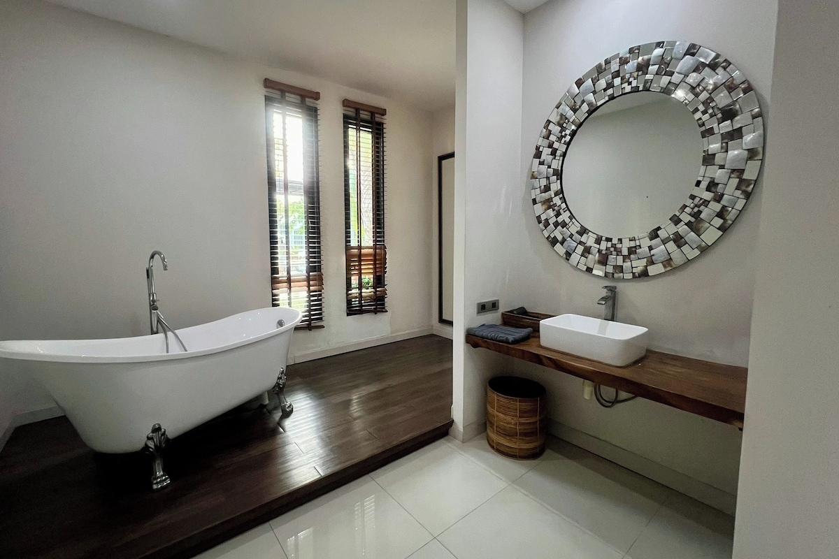 Un baño moderno con una bañera independiente, un espejo redondo con mosaicos, un mueble de tocador de madera con lavabo y una ventana con persianas.