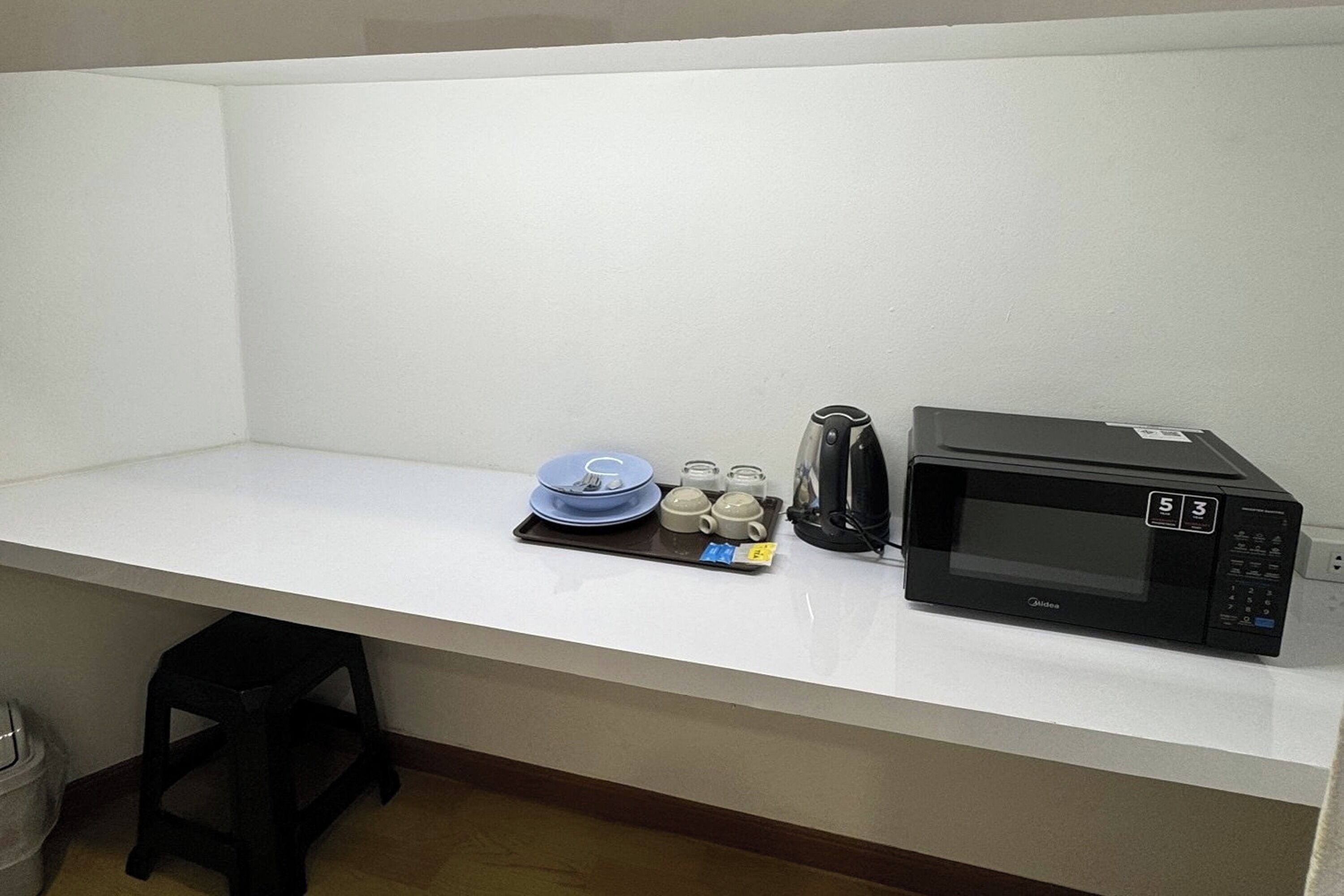 Een kitchenette met een magnetron, een koffiezetapparaat, een schaal met servies en een klein krukje.
