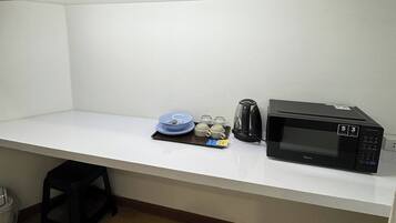 Een kitchenette met een magnetron, een koffiezetapparaat, een schaal met servies en een klein krukje.