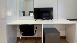 Quarto moderno e minimalista com uma secretária branca, uma cadeira preta, uma televisão de ecrã plano, um espelho e um pequeno móvel cinzento para arrumação.