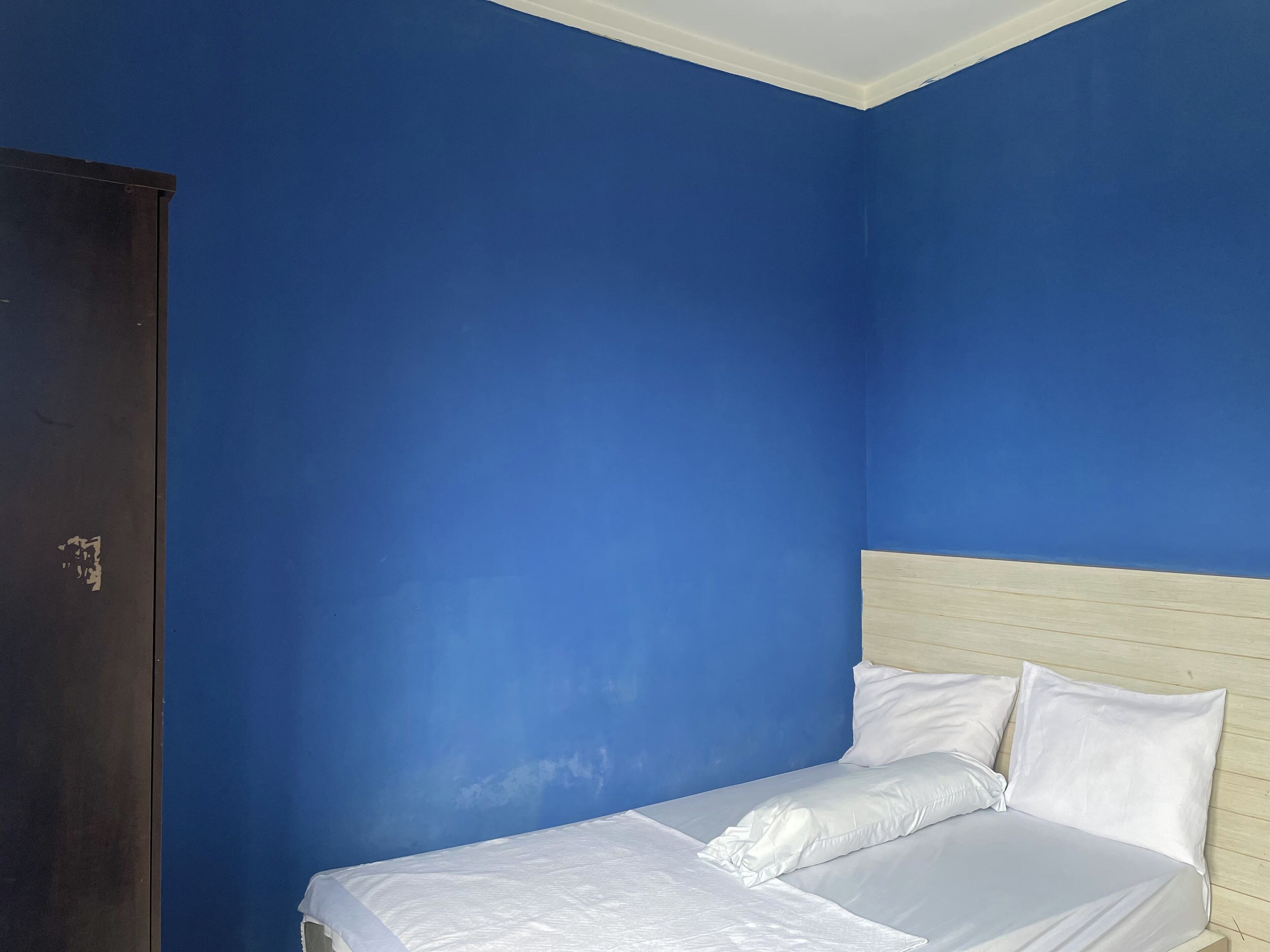 Un lit simple avec une literie blanche, dans une pièce aux murs bleus et dotée d’une tête de lit en bois.